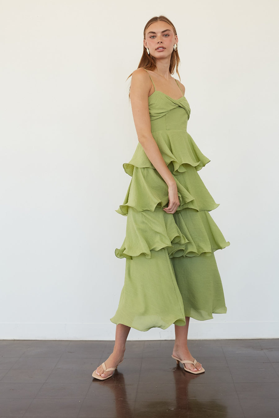 A woman models a light green tiered ruffle midi dress with a wrap-style bodice and wide-leg cropped pants, paired with clear strap heels, against a white studio wall.