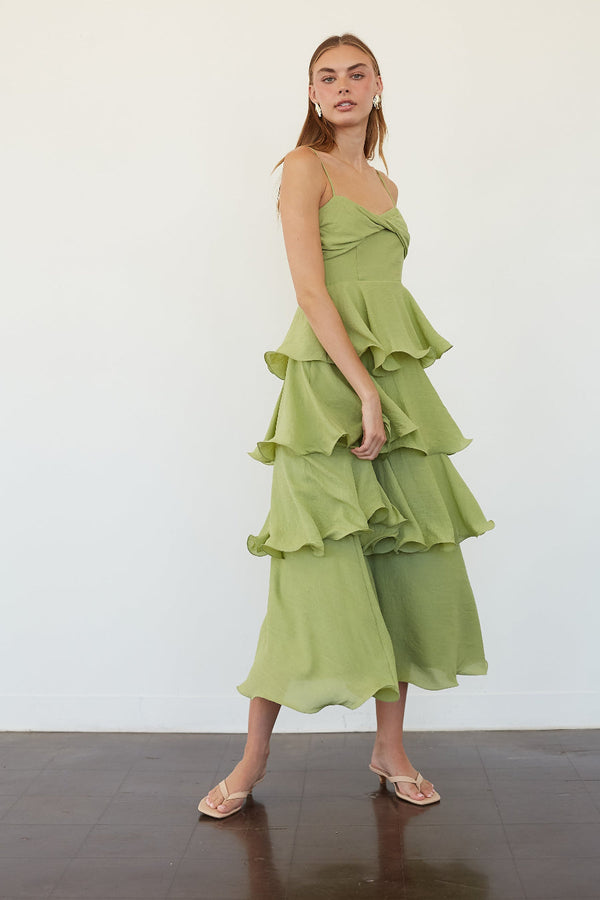 A woman models a light green tiered ruffle midi dress with a wrap-style bodice and wide-leg cropped pants, paired with clear strap heels, against a white studio wall.