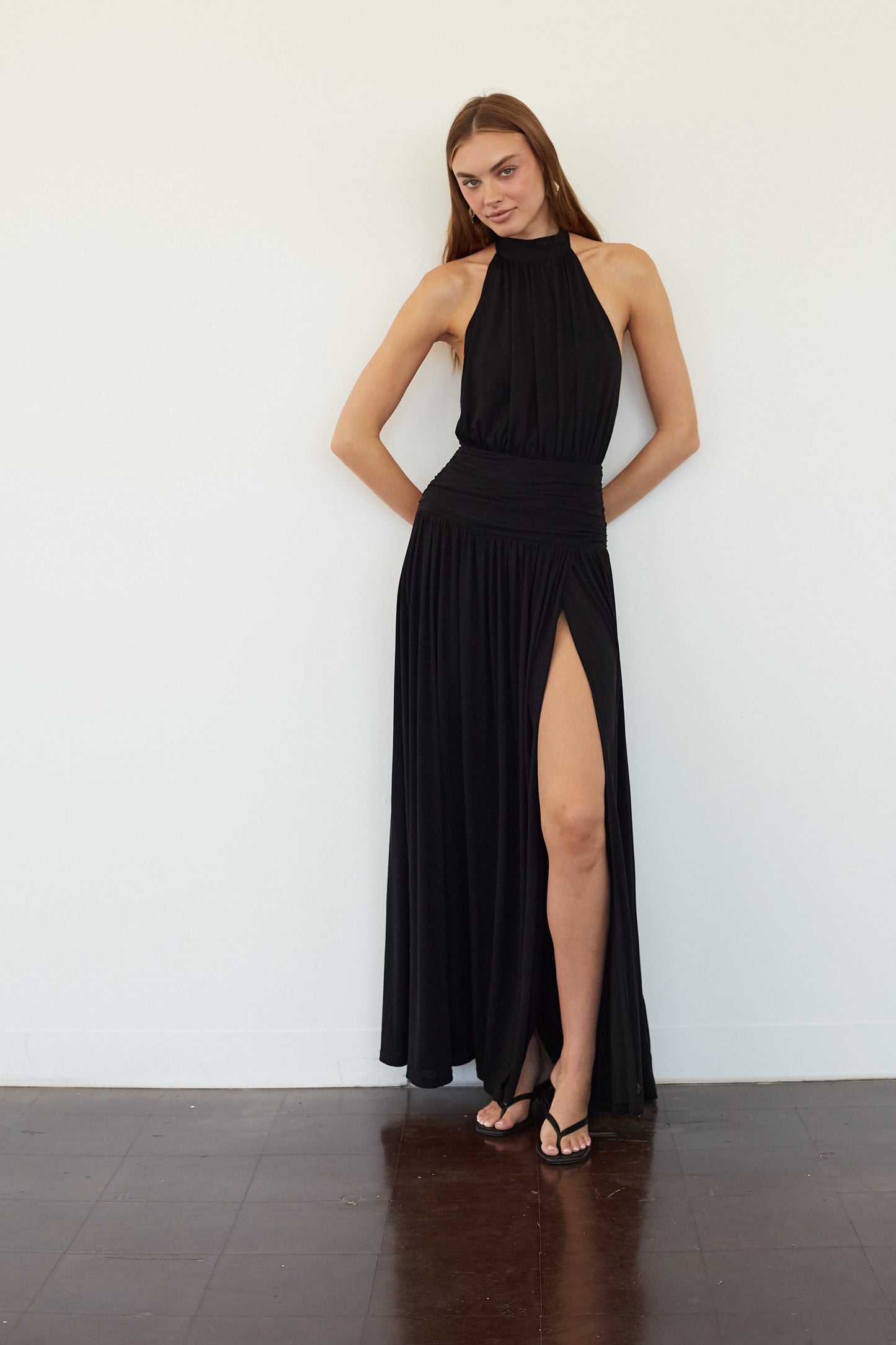 black-image | A woman modeling a sleeveless black halter-neck maxi dress with a high thigh slit, standing against a plain white wall on a dark wood floor.