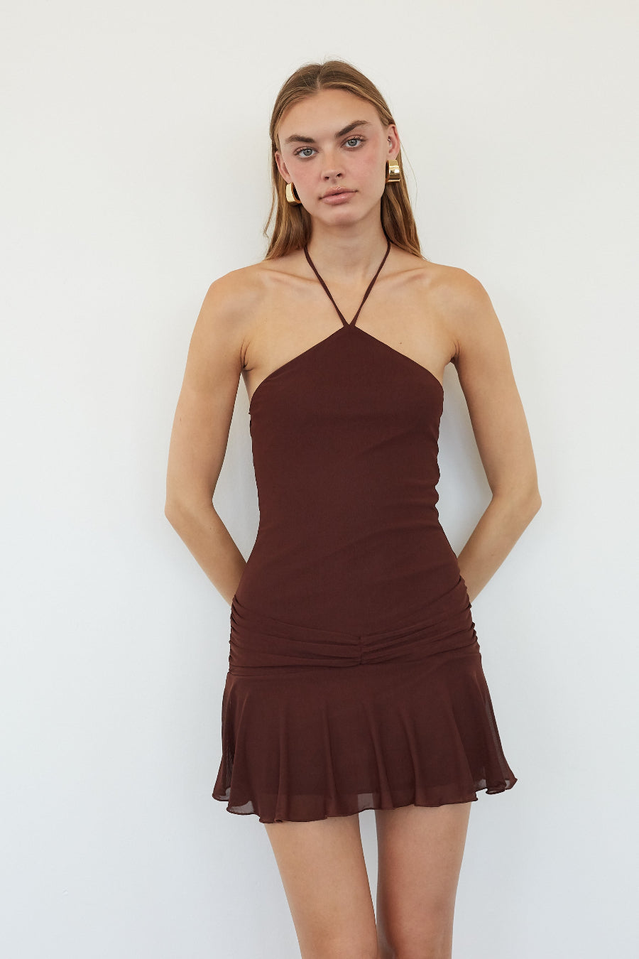 A woman modeling a dark brown halter-neck mini dress with a ruched bodice and flared ruffled hem, set against a plain white background.