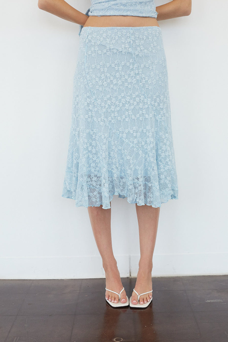 A light blue floral lace midi skirt worn by a model, paired with a matching light blue crop top and white strappy heels, standing against a white wall.