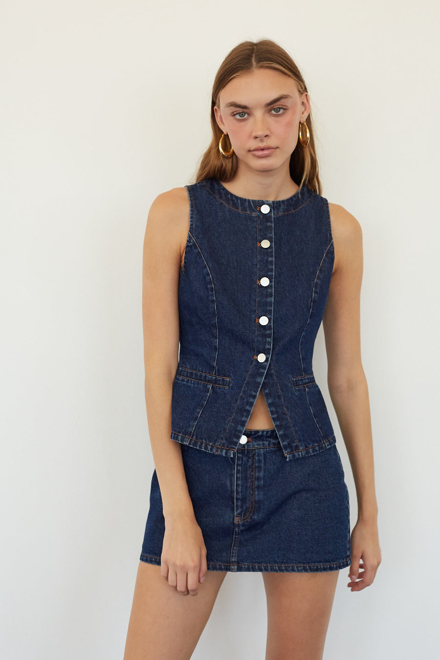 A model wearing a dark blue denim two-piece set consisting of a sleeveless button-front vest and a matching mini skirt, set against a plain white background.