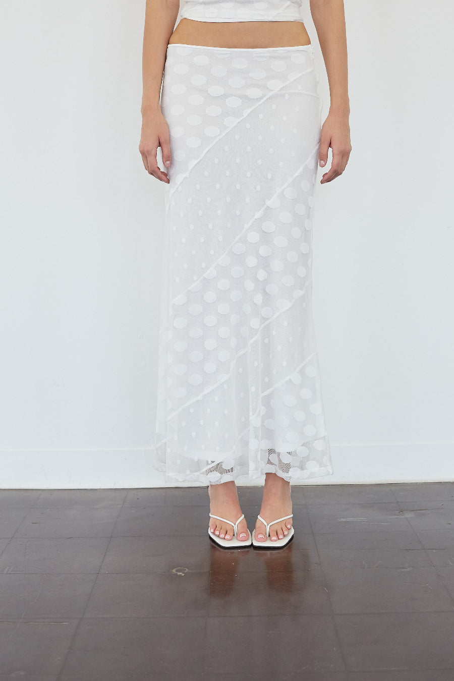 A model wearing a white sheer maxi skirt with a polka dot pattern, paired with white strappy sandals, standing against a white wall and dark wood floor.