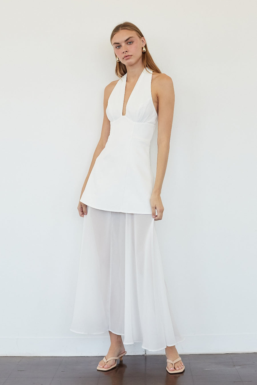 A woman modeling a white sleeveless halter-neck top with a flared waist and matching sheer white maxi skirt, paired with nude flat sandals, standing against a white wall.