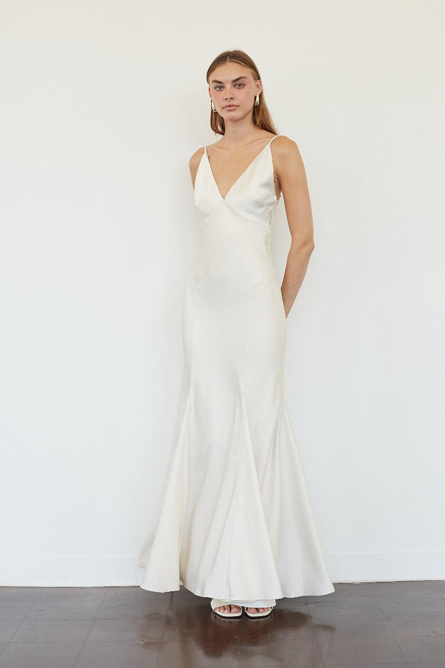 white-image | A woman modeling a floor-length ivory satin gown with a V-neckline and thin spaghetti straps, standing against a plain white wall.