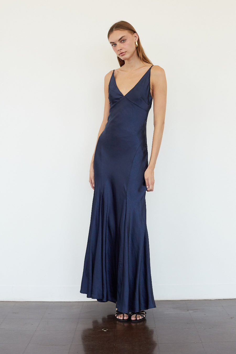 blue-image | A woman modeling a navy blue satin slip dress with a V-neckline and spaghetti straps, standing against a plain white wall.