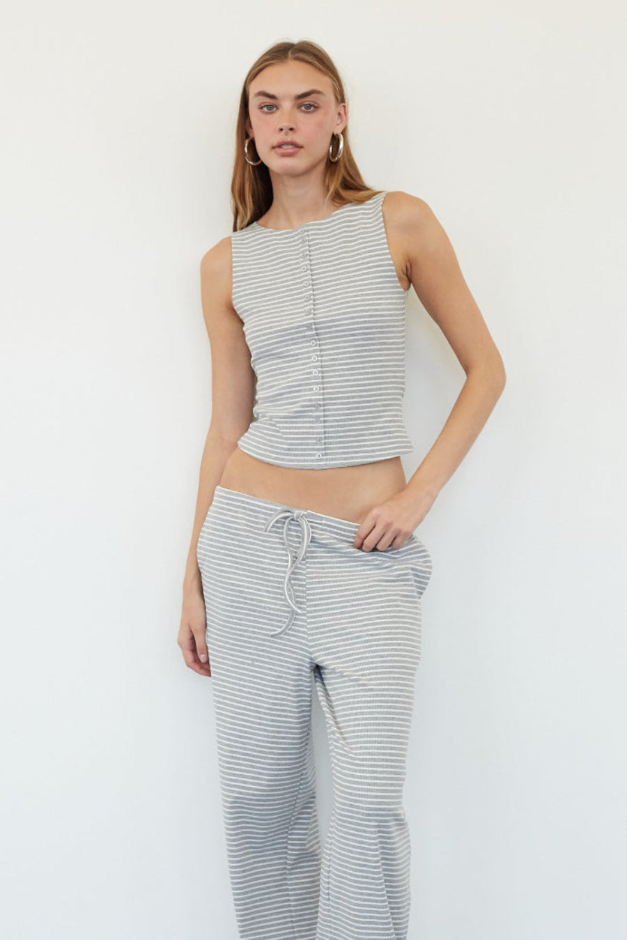 A woman modeling a matching two-piece loungewear set consisting of a sleeveless cropped tank top and wide-leg drawstring pants, both in a fine horizontal stripe pattern of gray and white, standing against a plain white wall.