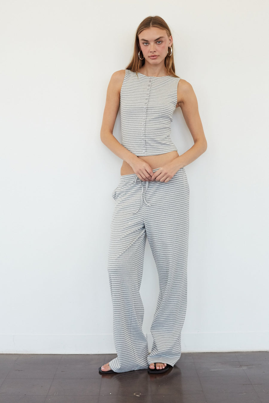 A woman modeling a matching two-piece loungewear set consisting of a sleeveless cropped top and wide-leg pants, both in a gray and white striped pattern, standing against a plain white wall.