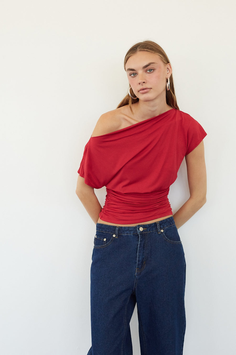 A woman modeling a red off-the-shoulder short-sleeve top with gathered draping, paired with dark blue jeans, against a plain white background.