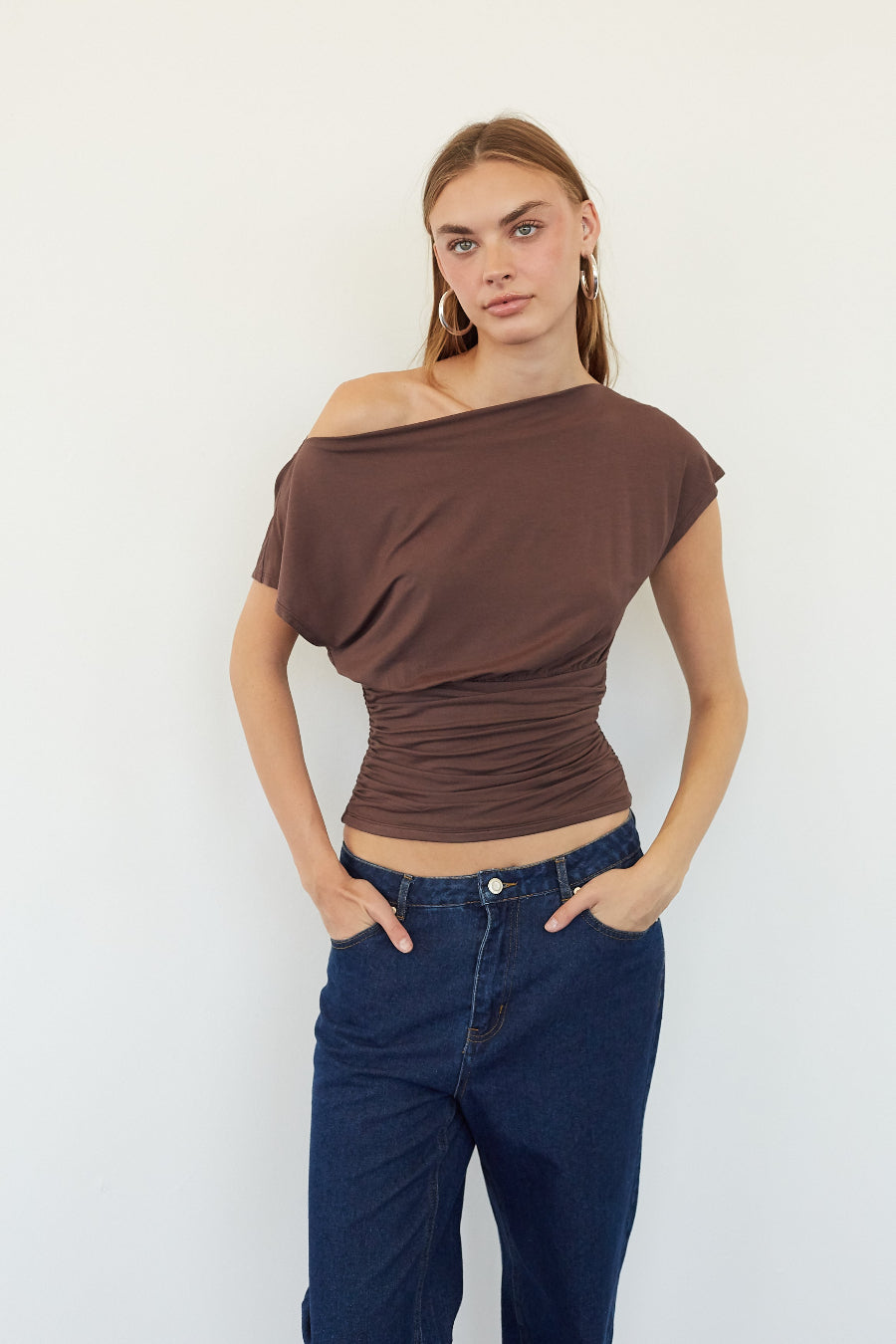 A woman modeling a brown off-the-shoulder ruched short-sleeve top paired with dark blue jeans, standing against a plain white wall.