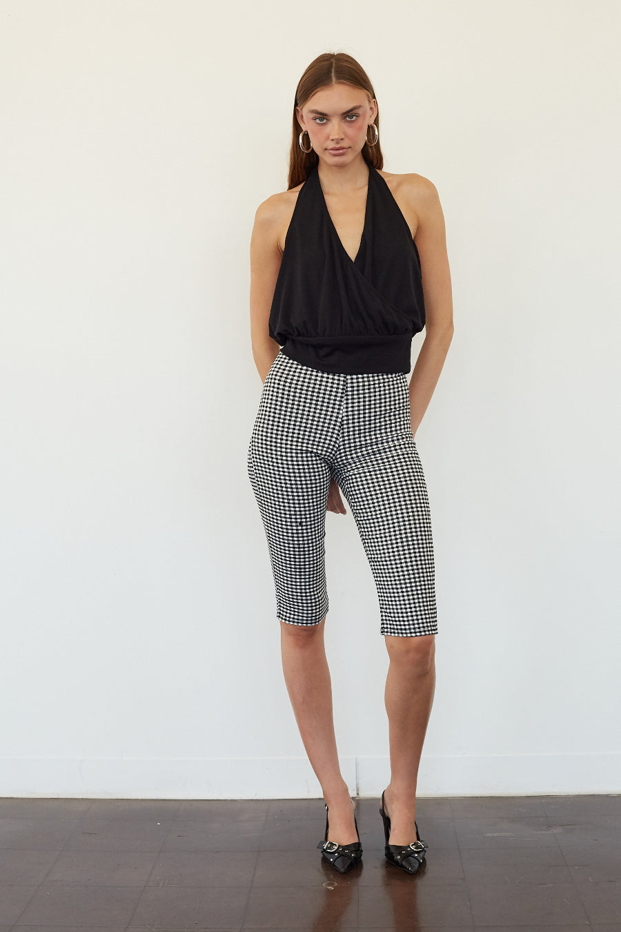 A woman stands against a plain white wall wearing a black halter top and black-and-white gingham capri pants, paired with black pointed heels.