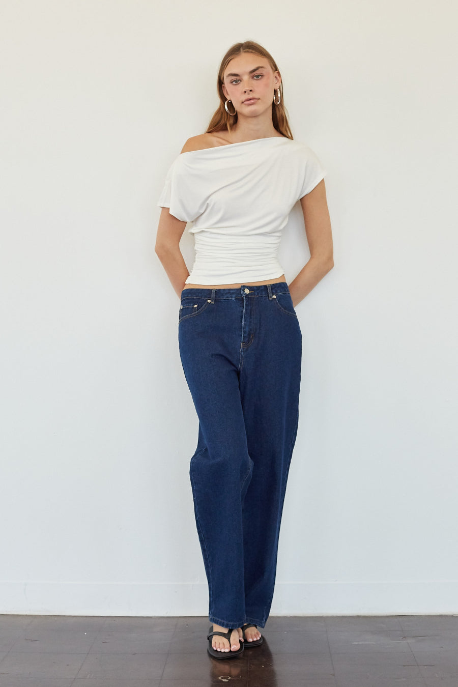 A woman stands against a plain white wall wearing a white off-the-shoulder short-sleeve crop top, dark blue straight-leg jeans, and black sandals.