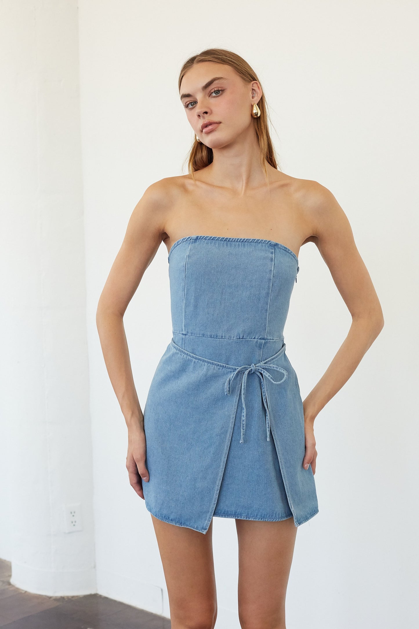 A woman modeling a light blue denim strapless mini dress with a tie waist, standing against a white wall.