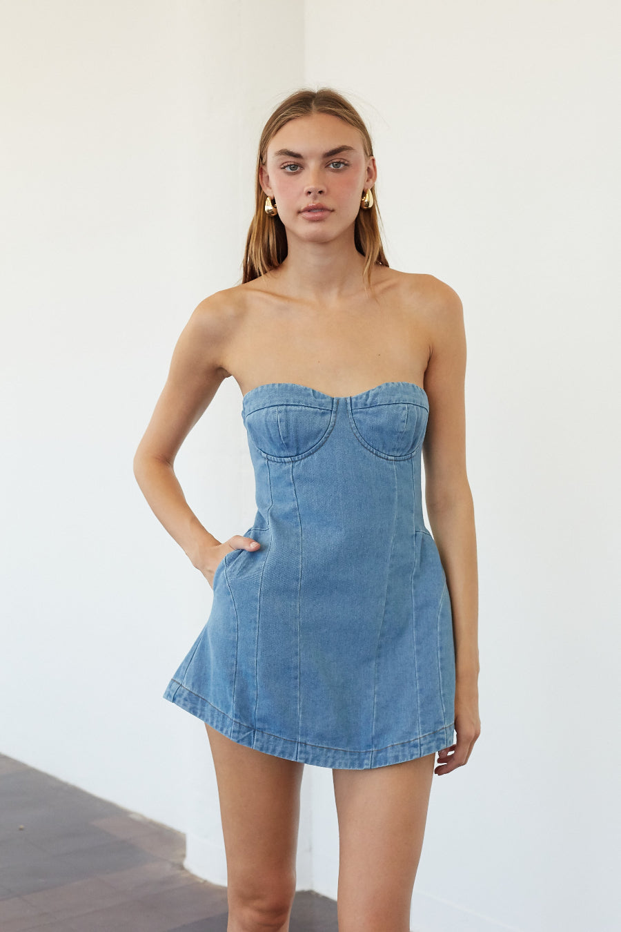 A woman modeling a strapless light blue denim mini dress with a sweetheart neckline and a flared skirt, standing against a plain white wall.