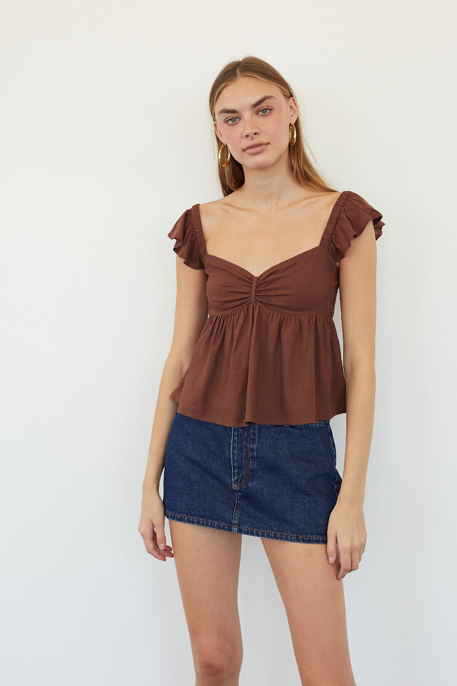 A woman modeling a brown ruffle-sleeve peplum top with a gathered sweetheart neckline, paired with a blue denim mini skirt, against a plain white studio background.
