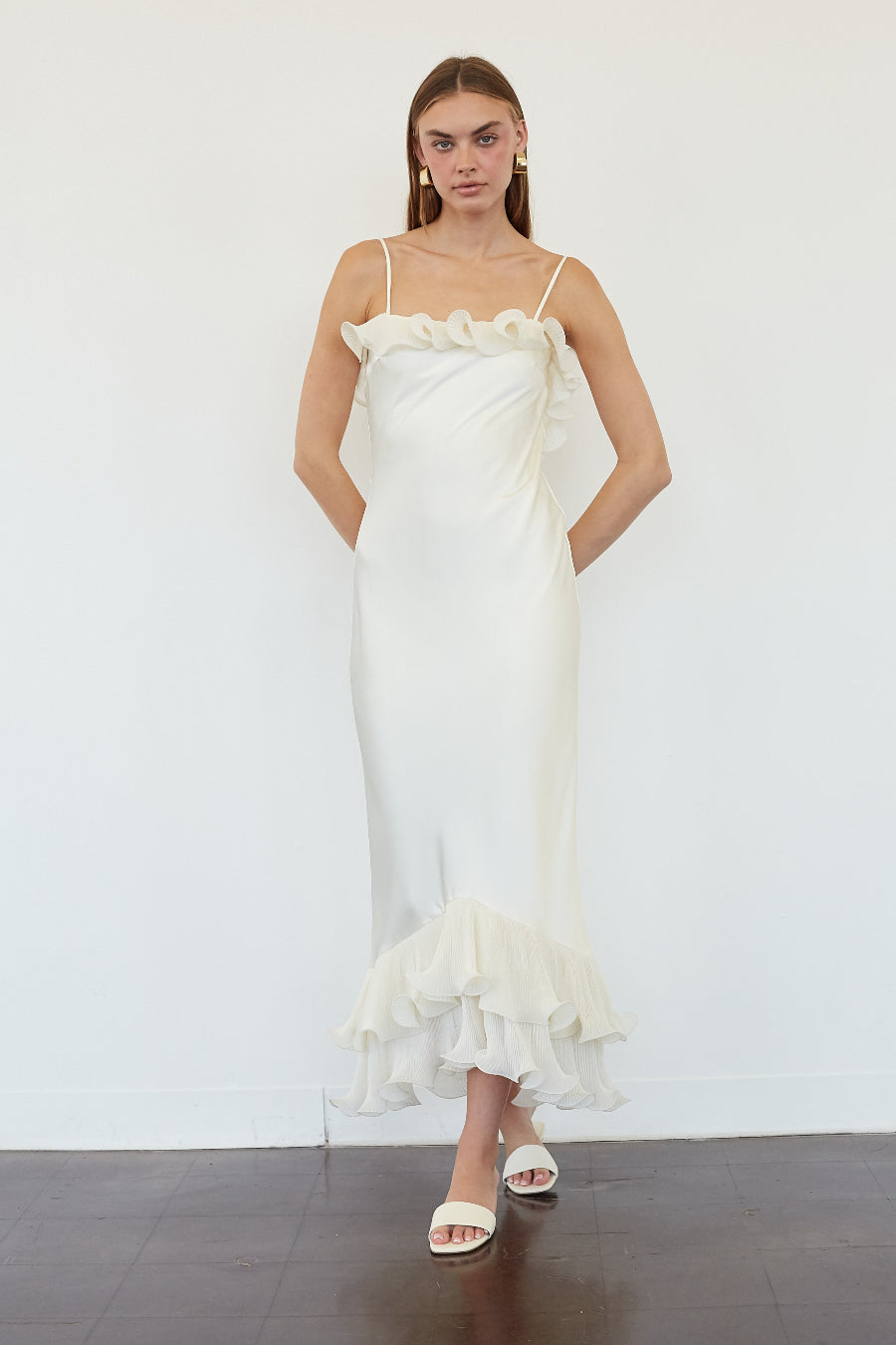 A woman modeling a white ruffled spaghetti-strap maxi dress with a fitted silhouette and a layered ruffle hem, standing against a white wall.