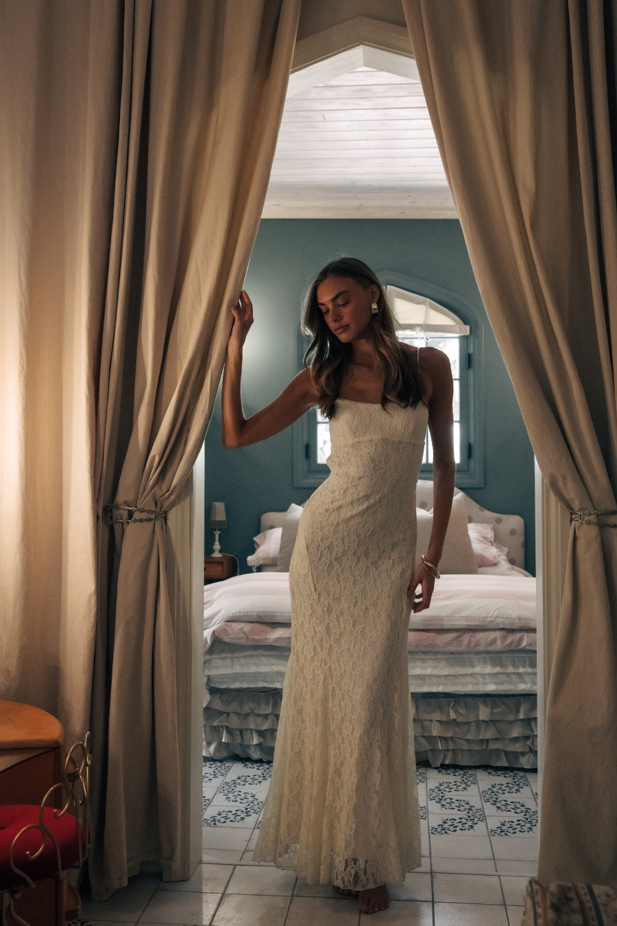 Woman in a white dress standing in a bedroom with curtains drawn back.