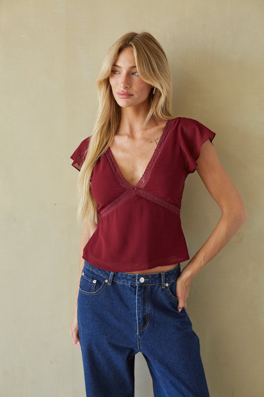 A woman modeling a burgundy short-sleeve top with a deep V-neck and lace trim, paired with blue jeans, standing against a textured beige wall.