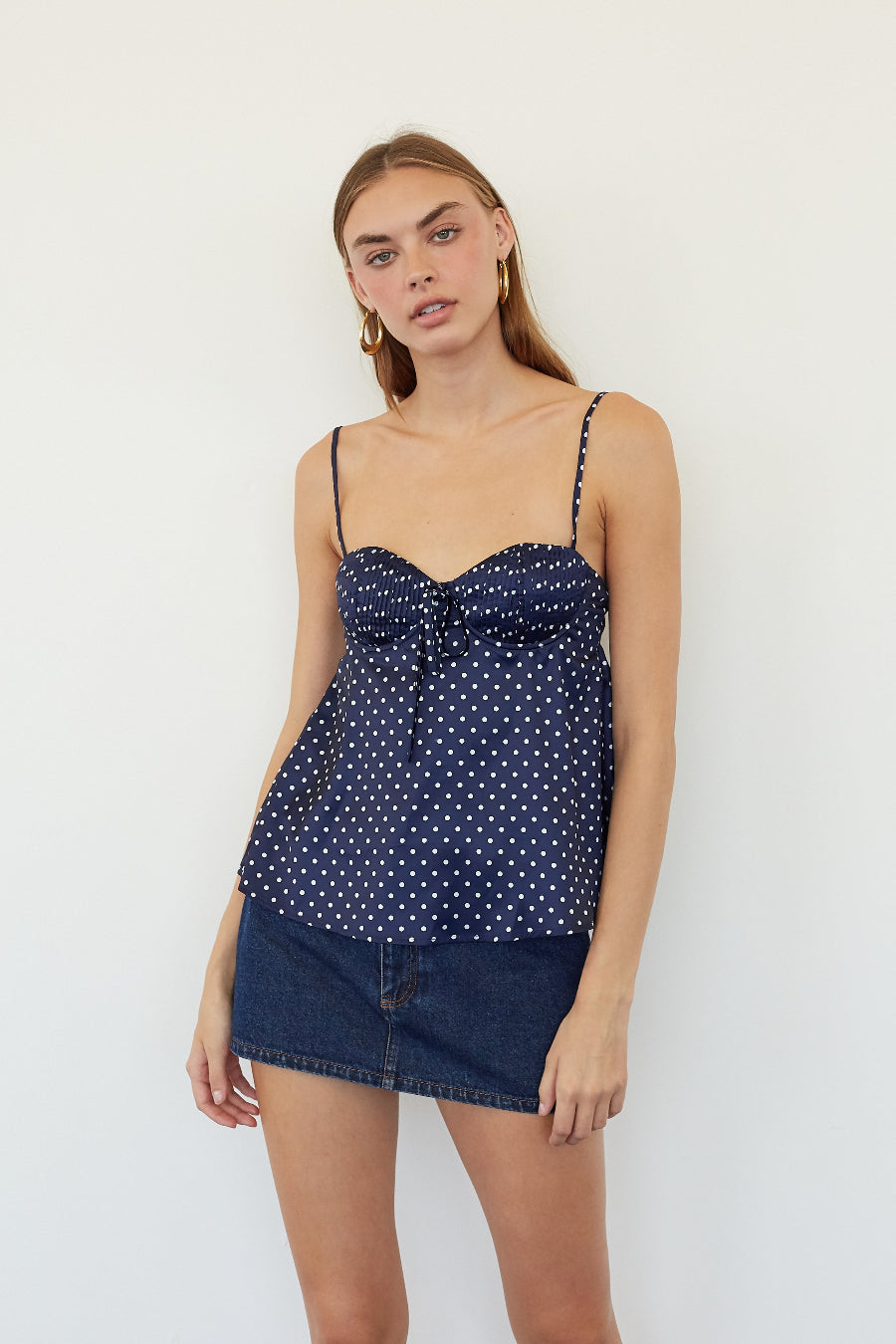 A woman modeling a navy blue camisole top with white polka dots and thin spaghetti straps, paired with a dark denim mini skirt, against a plain white studio background.