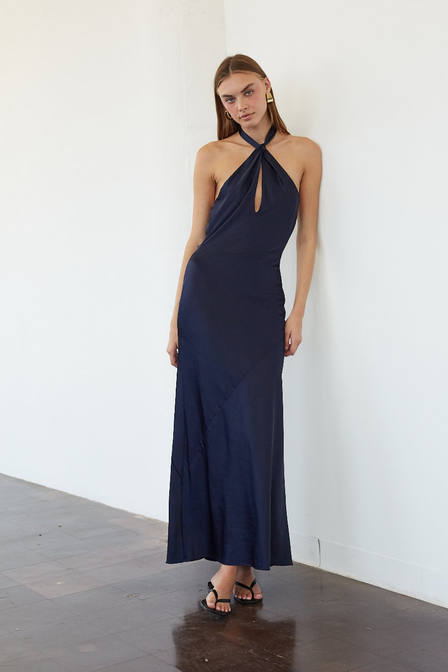 A woman modeling a navy blue halter-neck maxi dress with a keyhole cutout at the chest, standing against a white wall on a dark wood floor.