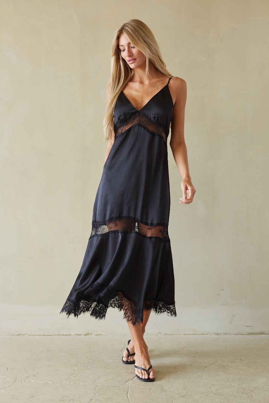 black-image | A woman modeling a dark navy blue maxi slip dress with sheer lace trim at the bust and hem, paired with black flip-flop sandals against a neutral beige wall.