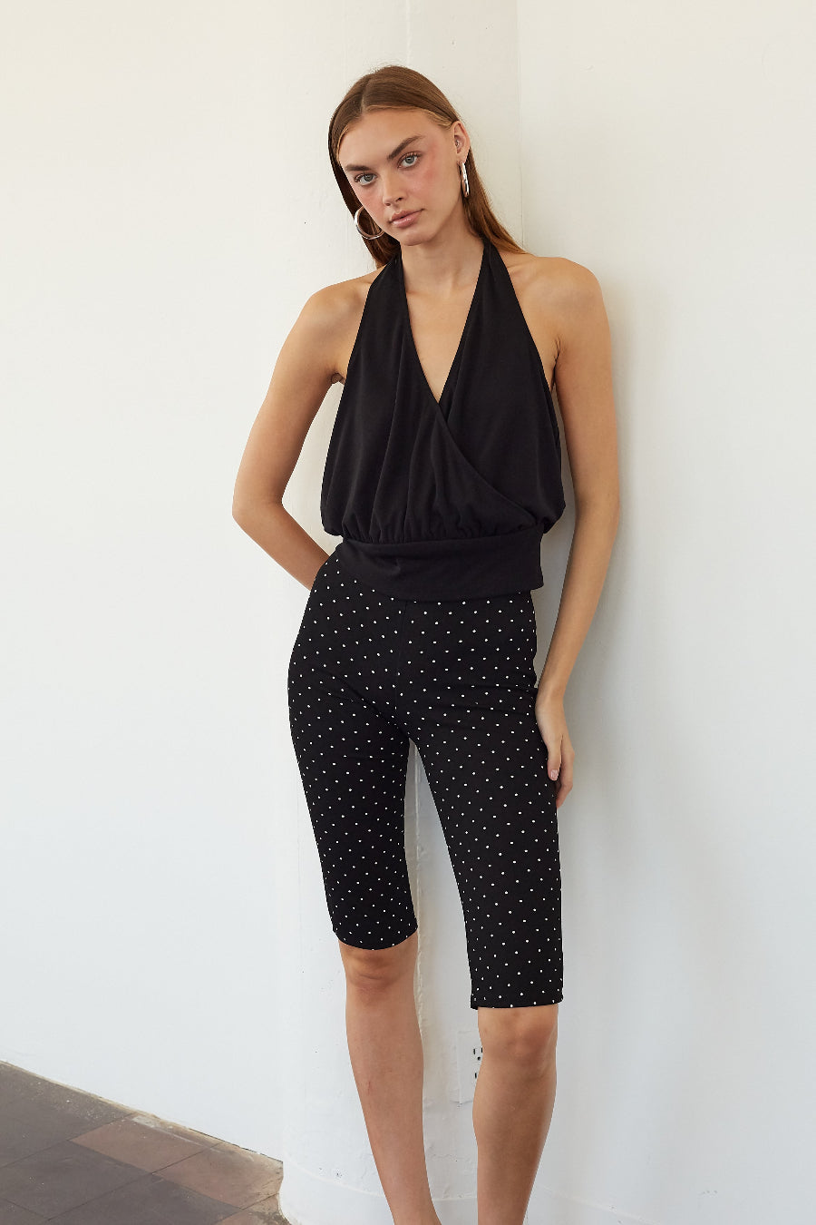 A woman modeling a black halter top and black capri pants with a small white polka dot pattern, standing against a plain white wall.