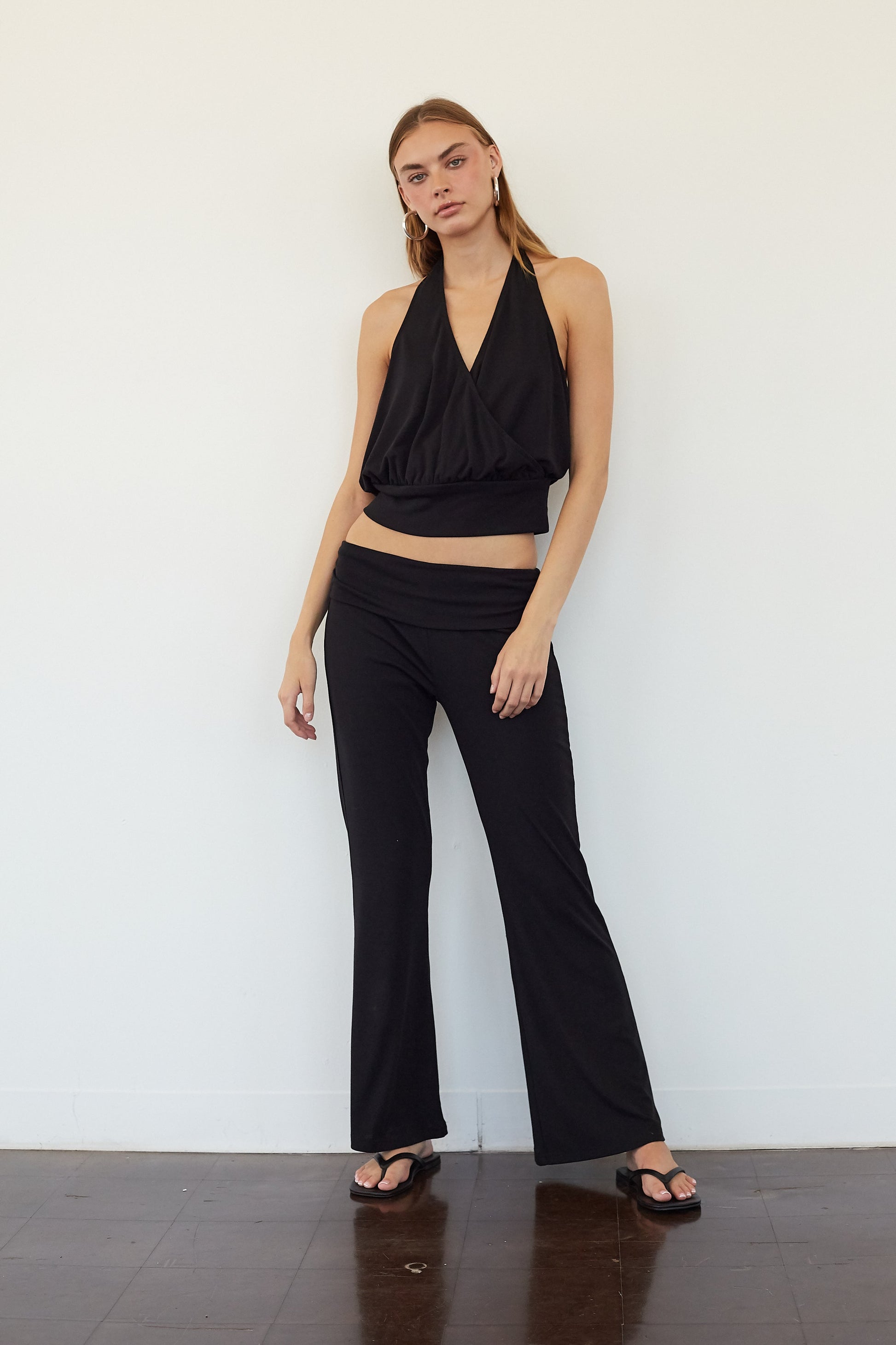 A woman modeling a black two-piece outfit consisting of a halter crop top and matching wide-leg pants, standing against a plain white wall on a dark wood floor.