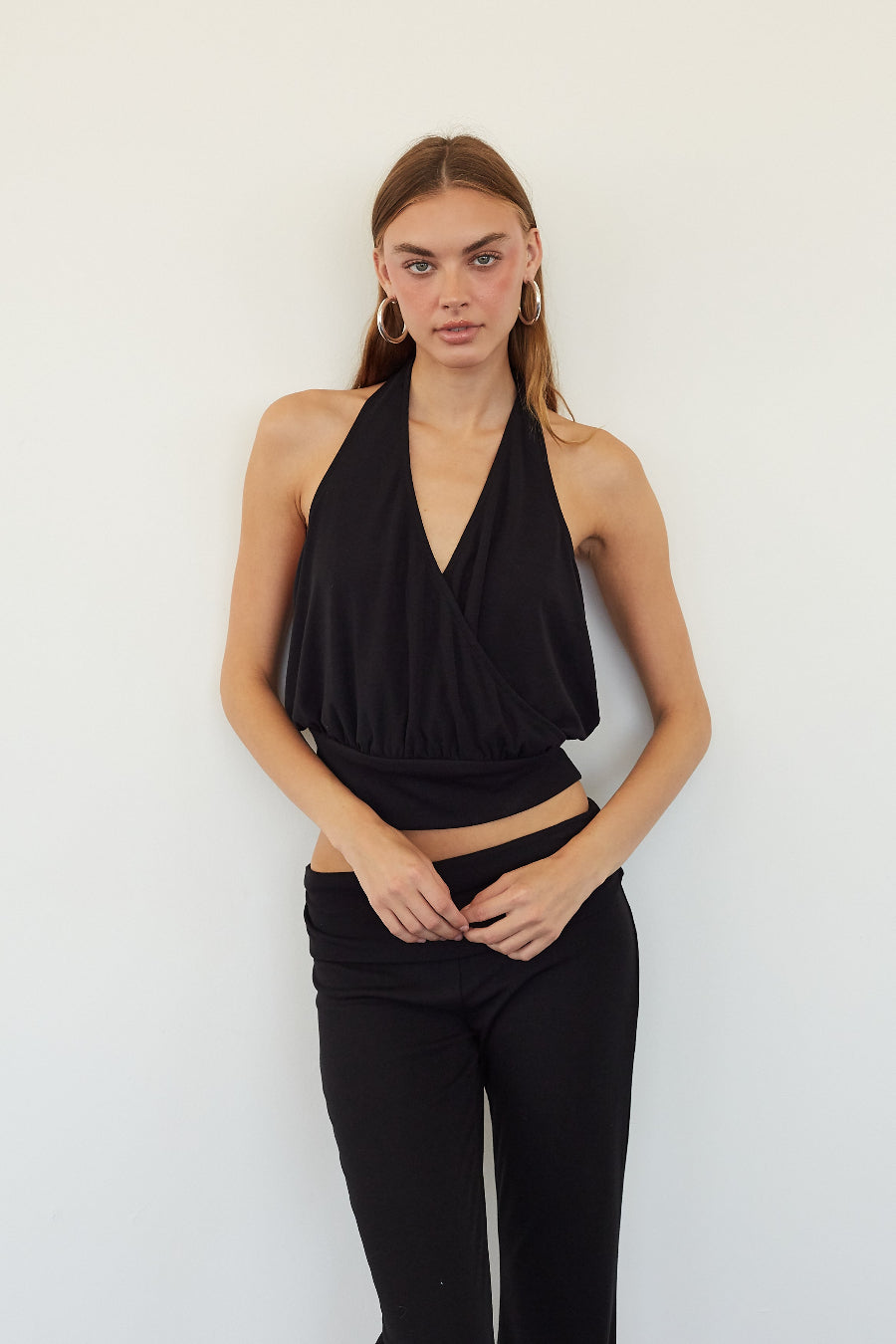 A woman modeling a black halter wrap-style crop top and black pants against a plain white wall.